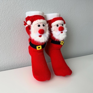 May include: A pair of bright red knit socks featuring a three-dimensional Santa Claus face on each. The Santa design includes a white faux fur beard, a red nose, black eyes, and a black belt with a yellow buckle around the ankle area.