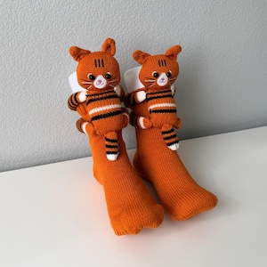 May include: A pair of bright orange knit socks, each featuring a plush, three-dimensional orange cat with black stripes attached to the cuff. The cat figures have white muzzles, embroidered whiskers, and small tails that hang down the front of the socks.