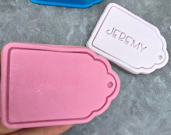 May include: Two cookie cutters in the shape of a tag. One is pink and the other is blue. The blue cutter is a mold for the pink cutter. The pink cutter has the name "JEREMY" embossed on it.