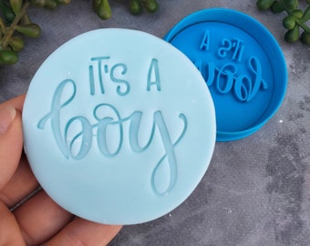 May include: A blue silicone cookie cutter with the text "It's a boy" in a cursive font. The cookie cutter is on a light blue background.
