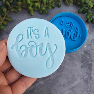 May include: A blue silicone cookie cutter with the text "It's a boy" in a cursive font. The cookie cutter is on a light blue background.