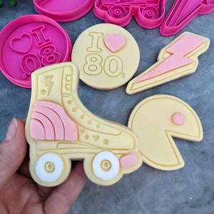 May include: A set of pink and yellow cookie cutters featuring a roller skate, lightning bolt, Pac-Man, and "I love the 80s" design.