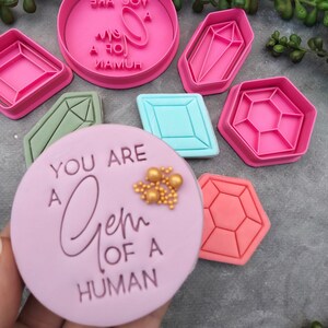 May include: A set of pink and green plastic cookie cutters shaped like different gemstones. The cutters are arranged on a grey surface. A purple cookie is in the centre of the image with gold sprinkles and the text "You are a Gem of a Human".