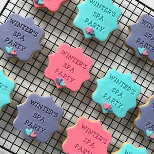 Op de afbeelding: Een close-up van negen gedecoreerde suikerkoekjes met de tekst "Winter's Spa Party" op elk koekje. De koekjes zijn versierd met roze, blauwe en paarse glazuur en hebben kleine hartvormige decoraties.