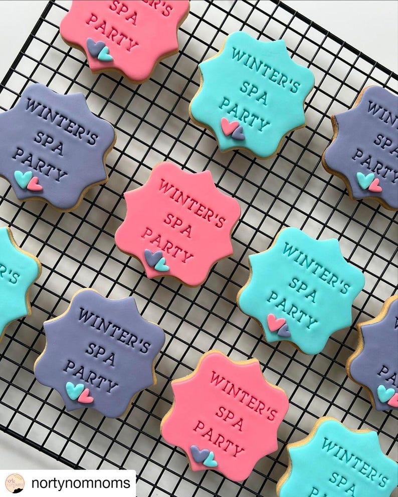 Op de afbeelding: Een close-up van negen gedecoreerde suikerkoekjes met de tekst "Winter's Spa Party" op elk koekje. De koekjes zijn versierd met roze, blauwe en paarse glazuur en hebben kleine hartvormige decoraties.