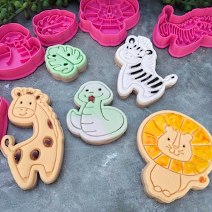 Op de afbeelding: Een set van negen koekjesvormen met verschillende dierenontwerpen, waaronder een giraffe, een leeuw, een slang, een zebra en een blad. De vormen zijn gemaakt van wit plastic en hebben een roze, gele en groene kleurstelling.