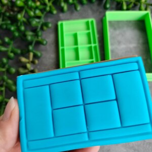 Op de afbeelding: Een blauwe rechthoekige koekjessnijder in de vorm van een tennisbaan, vastgehouden door een hand. Twee groene koekjessnijders staan op de achtergrond. De tennisbaan-snijder heeft een rechthoekige vorm met lijnen en vierkanten.
