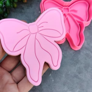 May include: Pink plastic cookie cutter in the shape of a bow. The cutter is open and the bow is visible.
