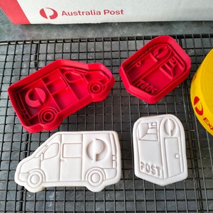 May include: Two red plastic cookie cutters shaped like a mail van and a post box. Next to them are two white cookies, one shaped like a mail van and the other shaped like a post box. The cookies are decorated with black icing and the text "POST".