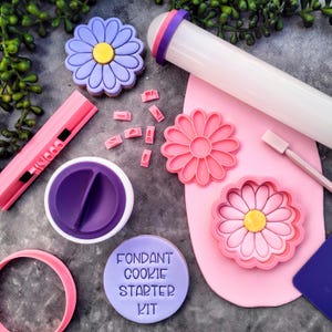 May include: A fondant cookie starter kit with a rolling pin, cutters, and a fondant smoother. The kit includes a daisy flower cutter, a daisy flower impression, and a round cutter. The kit also includes a set of letter stamps and a container of fondant.