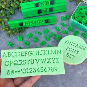 Op de afbeelding: Groene plastic alfabetstempels met een vintage lettertype. De stempels zitten in een doorzichtige plastic doos en bevatten hoofdletters, cijfers en symbolen. De tekst "Vintage Font Set" is gedrukt op een groene koek.