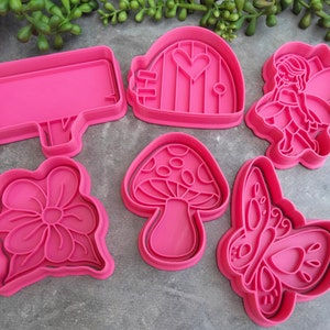 Op de afbeelding: Set van zes roze plastic koekjesvormen met een fee, een paddenstoel, een vlinder, een bloem, een huis en een bord.