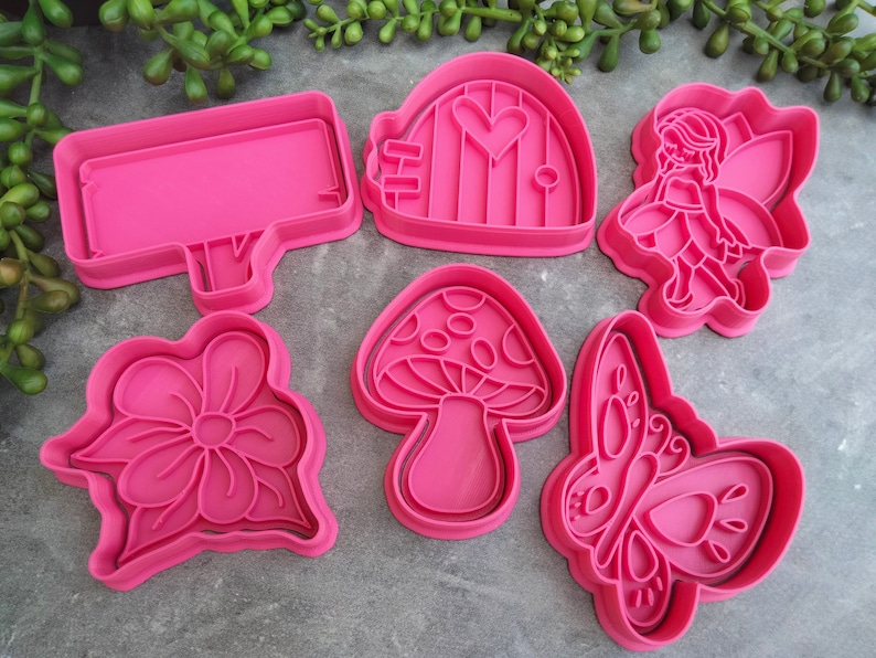 Op de afbeelding: Set van zes roze plastic koekjesvormen met een fee, een paddenstoel, een vlinder, een bloem, een huis en een bord.