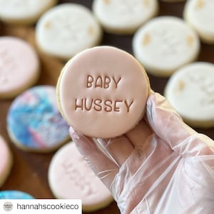 May include: A pink sugar cookie with the words "Baby Hussey" written in brown icing.