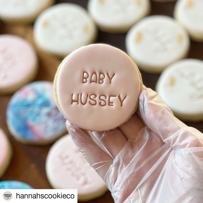 May include: A pink sugar cookie with the words "Baby Hussey" written in brown icing.