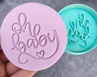May include: A set of two pink and turquoise silicone cookie cutters. The larger cutter is shaped like a circle and features the words "Oh Baby" in a cursive font with a small heart. The smaller cutter is also shaped like a circle and features the words "Oh Baby" in a cursive font with a small heart.