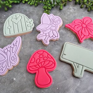 Op de afbeelding: Zes paarse en roze koekjesvormen in de vorm van een fee, een vlinder, een paddenstoel, een bloem, een deur en een blanco bord. De koekjesvormen liggen op een grijs oppervlak.