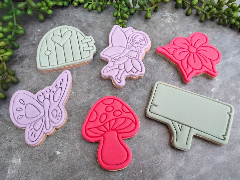 Op de afbeelding: Zes paarse en roze koekjesvormen in de vorm van een fee, een vlinder, een paddenstoel, een bloem, een deur en een blanco bord. De koekjesvormen liggen op een grijs oppervlak.