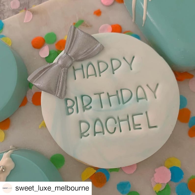 May include: A light blue round cake with white marbling and a silver bow on top. The cake is decorated with green lettering that says "Happy Birthday Rachel".