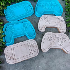 May include: A set of five cookie cutters shaped like video game controllers and handheld gaming devices. The cutters are made of blue and white plastic.