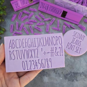 Op de afbeelding: Paarse alfabet- en cijferstempels met een "Skinny Jeans Font Set" label. De stempels zijn gemaakt van siliconen en zijn gerangschikt op een wit oppervlak.