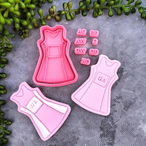 May include: Pink dress-shaped cookie cutters and stamps with the letters WA, GA, and other letter combinations. The cutters are in different sizes, and the stamps are for adding details. The items are arranged on a grey surface with green plant sprigs.
