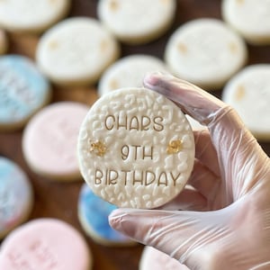May include: A white sugar cookie decorated with gold lettering that reads "Char's 9th Birthday". The cookie is being held by a gloved hand.