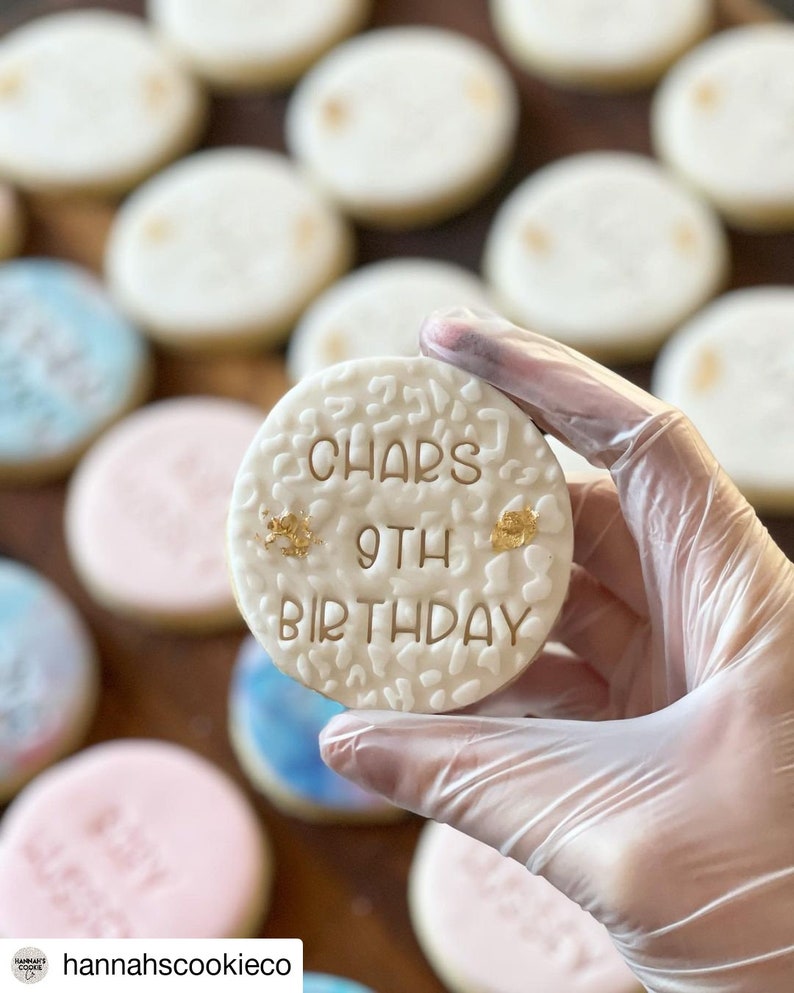 May include: A white sugar cookie decorated with gold lettering that reads "Char's 9th Birthday". The cookie is being held by a gloved hand.