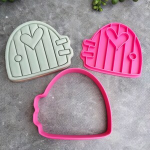 Op de afbeelding: Twee koekjesvormen in de vorm van een deur met een hart in het midden. Een vorm is lichtgroen en de andere is roze. Er is ook een roze omtrekkoekjesvorm.