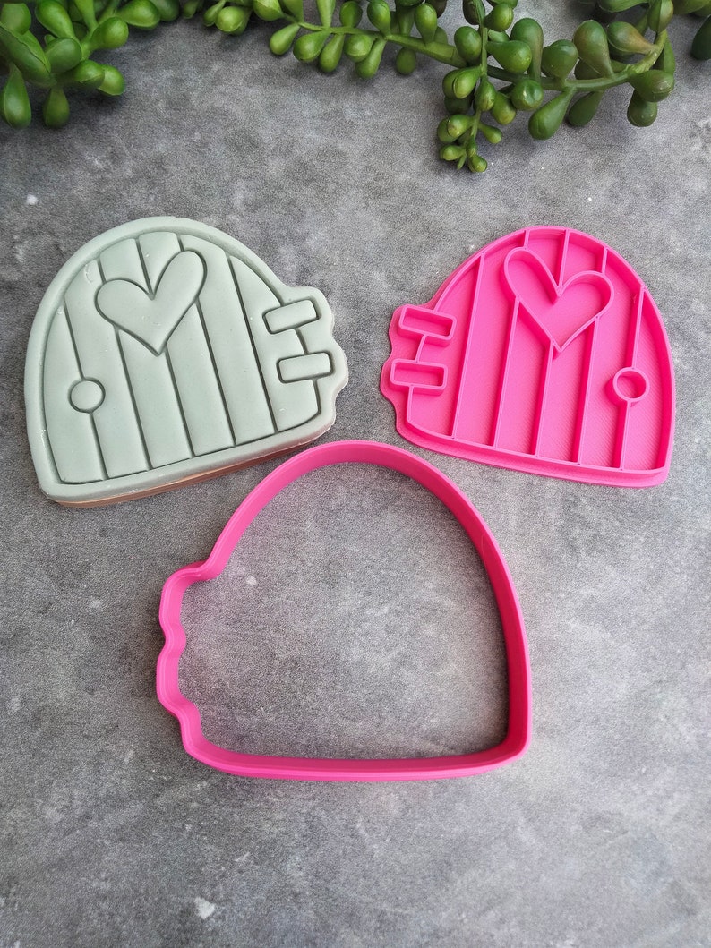 Op de afbeelding: Twee koekjesvormen in de vorm van een deur met een hart in het midden. Een vorm is lichtgroen en de andere is roze. Er is ook een roze omtrekkoekjesvorm.