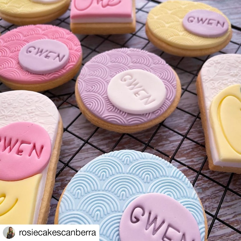 May include: A collection of decorated sugar cookies with a pastel color scheme. The cookies feature a variety of designs, including a floral pattern, a wave pattern, and a simple circle with the name "Gwen" written on it.