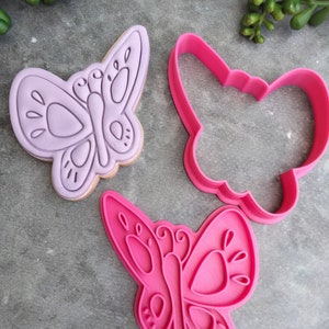 Op de afbeelding: Twee koekjesvormen in de vorm van vlinders, &eacute;&eacute;n paars en &eacute;&eacute;n roze. De paarse vorm is een gedetailleerd ontwerp met een vlinder vorm en de roze vorm is een eenvoudiger ontwerp.