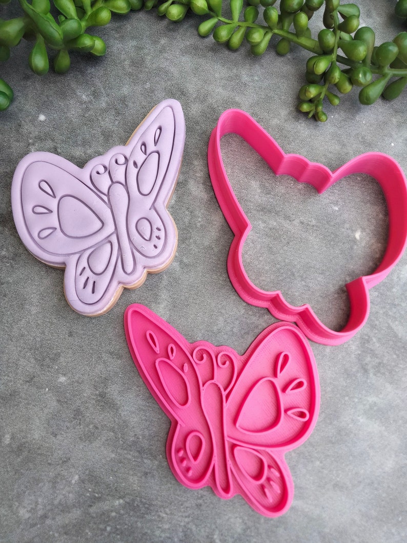Op de afbeelding: Twee koekjesvormen in de vorm van vlinders, &eacute;&eacute;n paars en &eacute;&eacute;n roze. De paarse vorm is een gedetailleerd ontwerp met een vlinder vorm en de roze vorm is een eenvoudiger ontwerp.