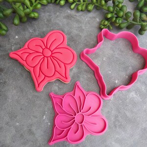 Op de afbeelding: Drie roze plastic koekjesvormen in de vorm van bloemen. De vormen liggen op een grijs oppervlak.