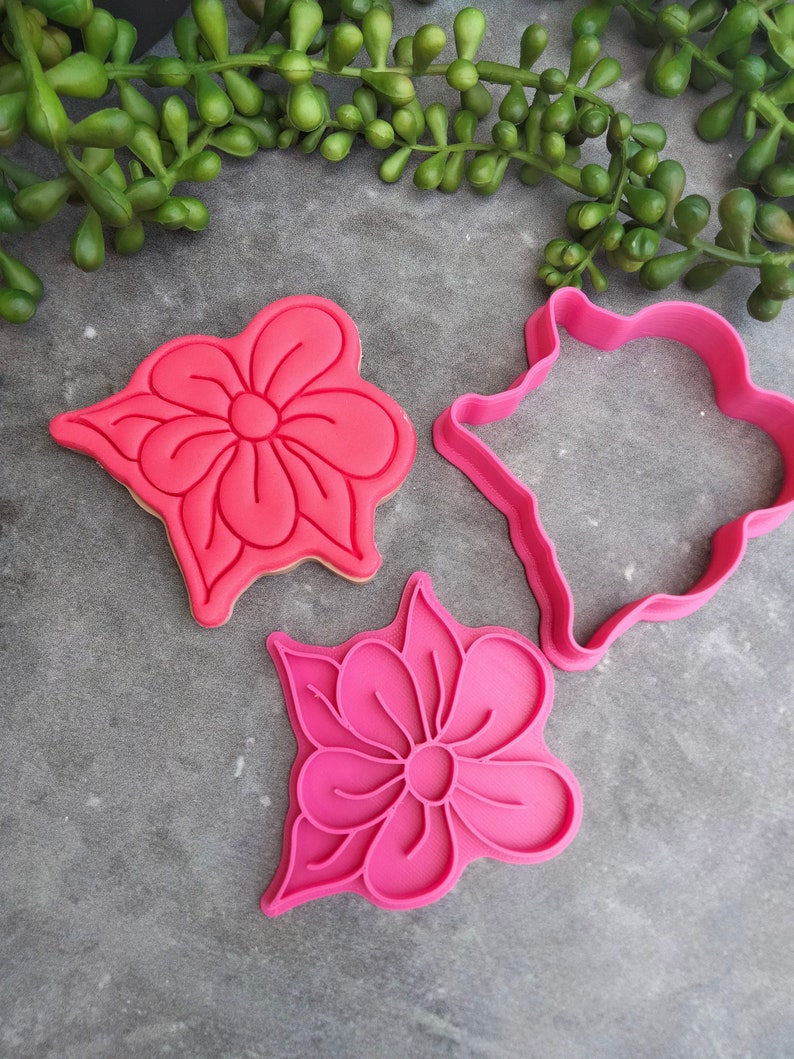 Op de afbeelding: Drie roze plastic koekjesvormen in de vorm van bloemen. De vormen liggen op een grijs oppervlak.