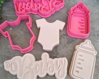 May include: Set of five pink and white plastic cookie cutters. The cutters are shaped like a baby onesie, a baby bottle, and the word "Baby" twice.