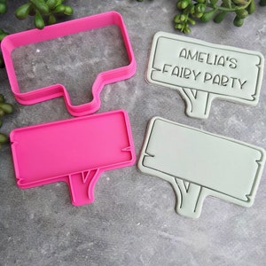 Op de afbeelding: Twee roze en grijze plastic koekjesvormen in de vorm van borden met een boomstam aan de onderkant. De grotere vorm heeft de tekst "Amelia's Fairy Party" erop.
