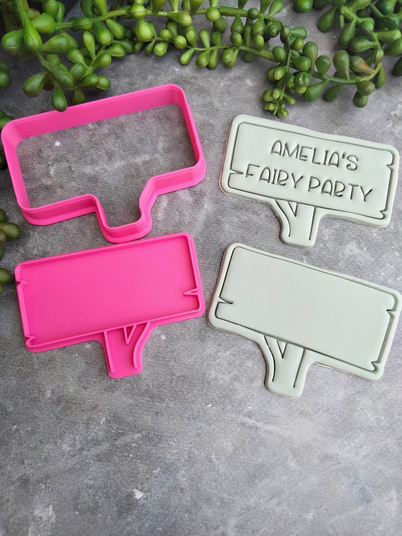 Op de afbeelding: Twee roze en grijze plastic koekjesvormen in de vorm van borden met een boomstam aan de onderkant. De grotere vorm heeft de tekst "Amelia's Fairy Party" erop.