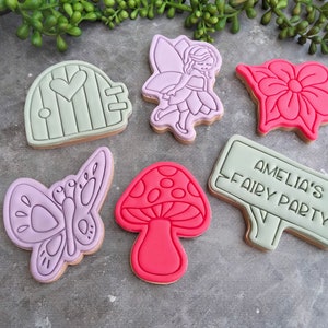 Op de afbeelding: Een set van zes gedecoreerde suikerkoekjes met een fee, een vlinder, een paddenstoel, een bloem, een deur en een bord met de tekst "Amelia's Fairy Party".