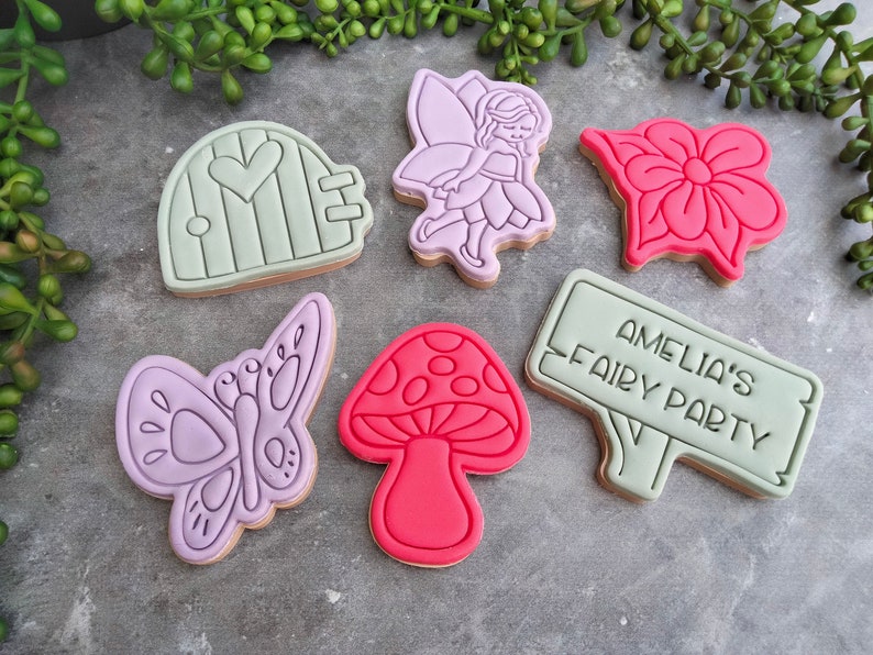 Op de afbeelding: Een set van zes gedecoreerde suikerkoekjes met een fee, een vlinder, een paddenstoel, een bloem, een deur en een bord met de tekst "Amelia's Fairy Party".