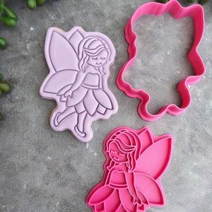Op de afbeelding: Twee roze plastic koekjesvormen in de vorm van een fee. E&eacute;n vorm is massief en de andere is een omtrek.