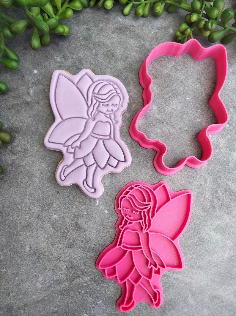 Op de afbeelding: Twee roze plastic koekjesvormen in de vorm van een fee. E&eacute;n vorm is massief en de andere is een omtrek.