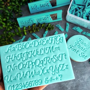 May include: A turquoise blue silicone stamp set with a cursive font alphabet, numbers, and symbols. The stamp set is labelled "Fancy Pants Font Set".