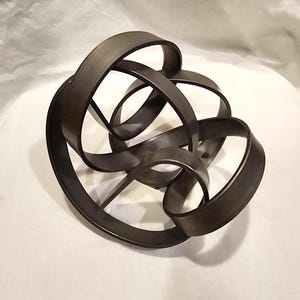Op de afbeelding: Een donkerbruin metalen sculptuur met een uniek, abstract ontwerp. Het sculptuur is gemaakt van meerdere in elkaar verweven lussen, waardoor een complexe en visueel interessante vorm ontstaat.