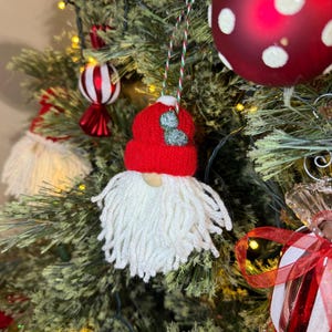May include: A gnome Christmas tree ornament featuring a bright red knitted hat with a small green leaf accent and a long, thick white yarn beard. The ornament hangs from a thin, twisted red, white, and green string against a pine tree.