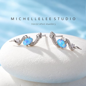 May include: A pair of silver-toned snake-shaped stud earrings featuring an oval-cut iridescent blue opal stone and a small clear accent stone. The text MICHELLELEE STUDIO Handcrafted Jewellery is visible above the earrings.