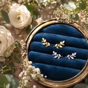 May include: Two pairs of leaf-shaped stud earrings, one pair in gold and one pair in silver, each featuring small white opal-like stones arranged along the branch. The earrings are displayed on a dark blue velvet insert within a round frame.