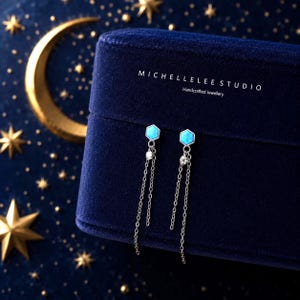 Puede incluir: Un par de pendientes plateados con un poste hexagonal de ópalo azul, un pequeño acento de cristal transparente y dos delicadas cadenas colgantes. Los pendientes se muestran sobre una caja de terciopelo azul oscuro con el texto MICHELLELEE STUDIO Handcrafted Jewellery.