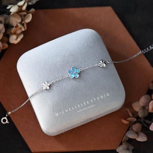 May include: A silver bracelet with three flower charms. The centre flower charm is blue and iridescent. The bracelet is in a grey velvet box with the text "MICHELLE LEE STUDIO handcrafted jewellery".