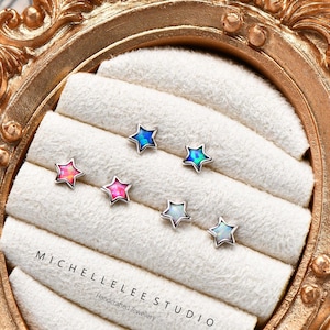 May include: A selection of star-shaped stud earrings in shades of pink, blue, and white, presented on a white velvet display. The earrings are showcased within a gold-toned ornate frame. The text "MICHELLELEE STUDIO" is visible on the display.